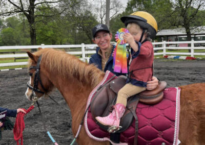 child-riding-pony-with-ribbon A young child wearing a riding helmet and pink boots sits on a brown pony with a maroon saddle pad, proudly holding a colorful ribbon. An adult stands beside them, smiling, with trees and a white fence in the background.