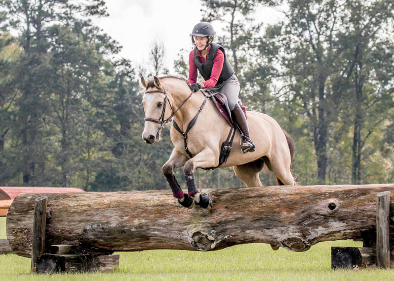 A rider wearing a helmet and protective vest is riding a light brown horse, jumping over a large wooden log obstacle outdoors with trees in the background.