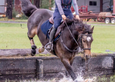 horse-jumping-water-obstacle A person wearing a helmet and protective vest rides a dark horse as it jumps into a shallow water obstacle, splashing water, with grass, trees, and a barn in the background.