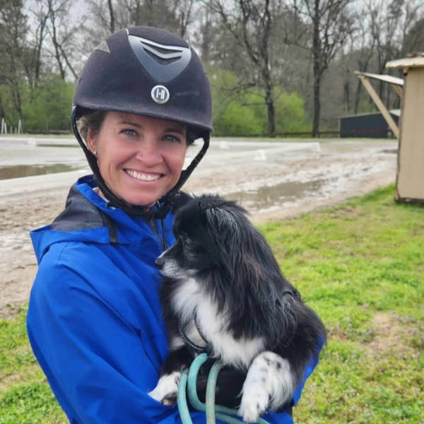 JulieNorman-TSDS2025 A person wearing a helmet and protective vest rides a dark horse as it jumps into a shallow water obstacle, splashing water, with grass, trees, and a barn in the background.